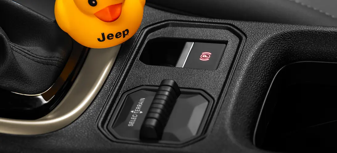 Freio de Estacionamento e Selec-Terrain Console central do Jeep Avenger com botão do freio de estacionamento elétrico, seletor Selec-Terrain e patinho amarelo de borracha com logotipo Jeep posicionado ao lado dos comandos.