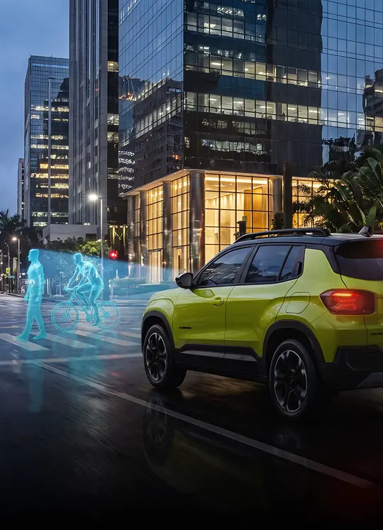 Jeep Avenger verde com parte traseira e lateral visíveis em avenida urbana entre edifícios iluminados, com elementos gráficos azuis sobre a faixa de pedestres.